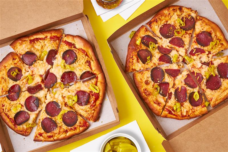 Two pizzas in boxes on a table with pickles.